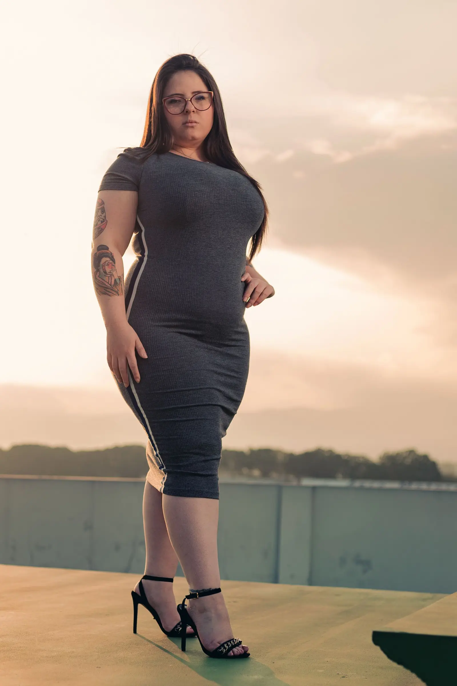 Woman in glasses and a fitted gray dress on a waterfront at sunset