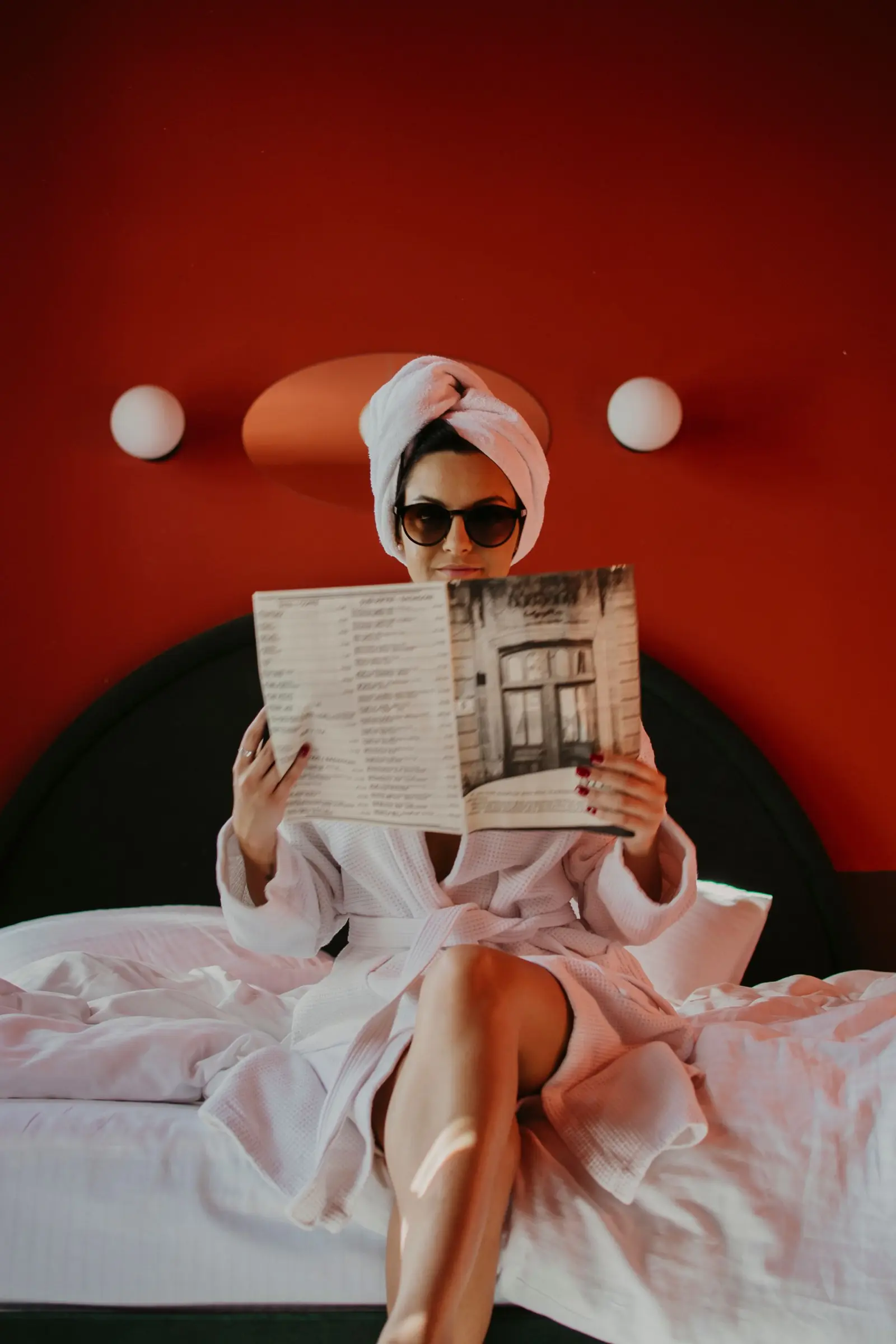 Woman in a robe and towel turban reading a magazine on a bed against a red wall