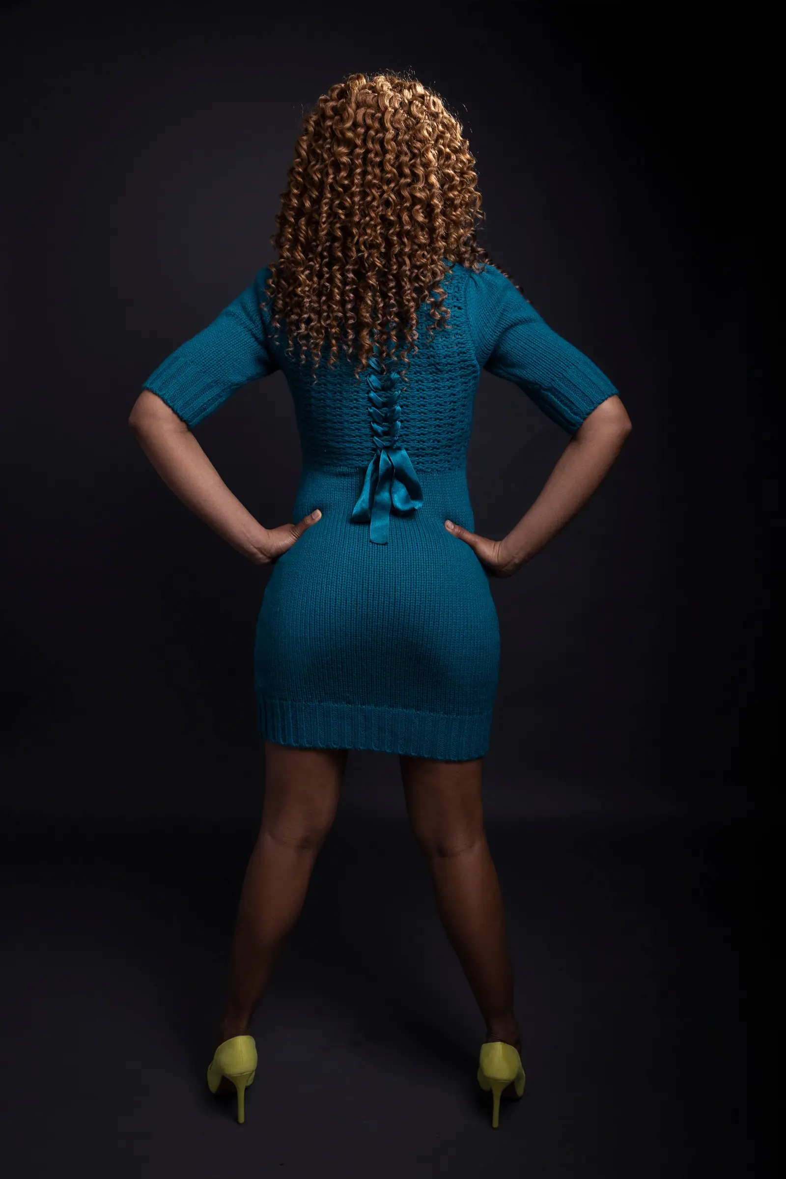 Woman in a fitted teal dress, seen from behind with hands on her hips